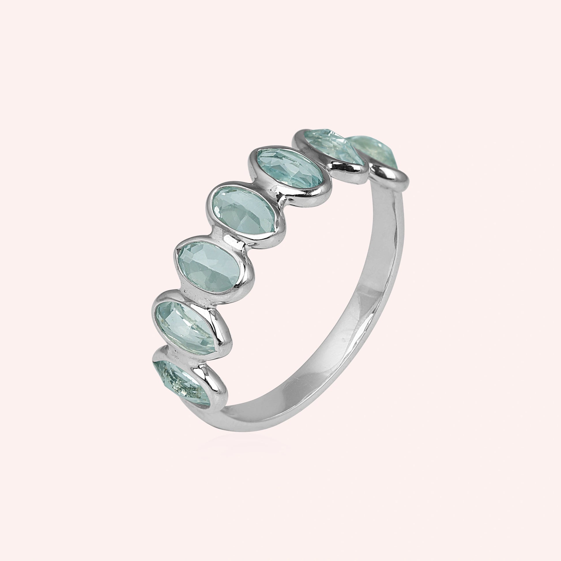 Sterling silver topaz ring band featuring seven bezel-set oval light blue gemstones on a plain pink background side angle.