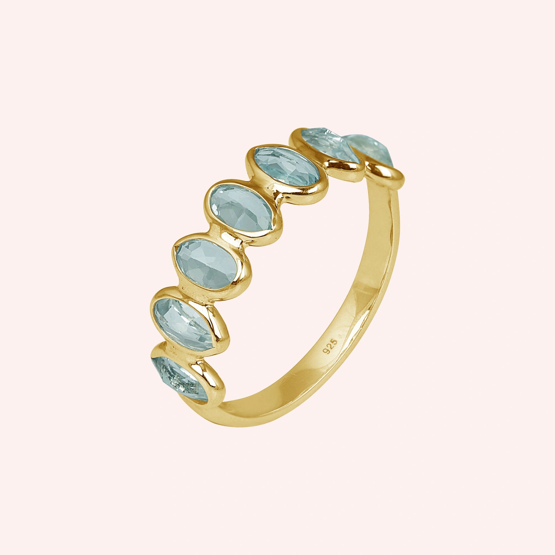 Gold-plated topaz ring band featuring seven bezel-set oval light blue gemstones on a plain pink background side angle.