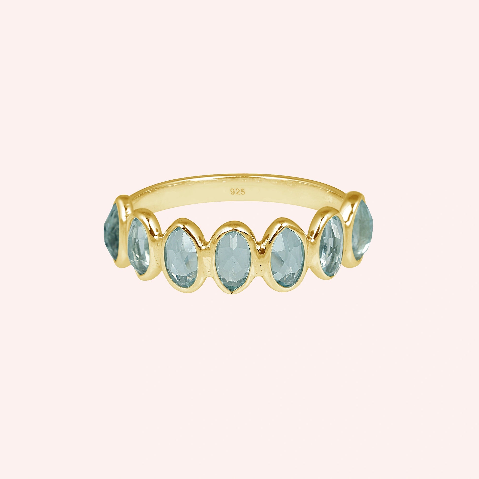 Gold-plated topaz ring band featuring seven bezel-set oval light blue gemstones on a pink background front view.