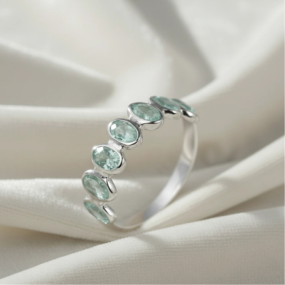 Sterling silver topaz ring band featuring seven bezel-set oval light blue gemstones on a white draped fabric slight angle.