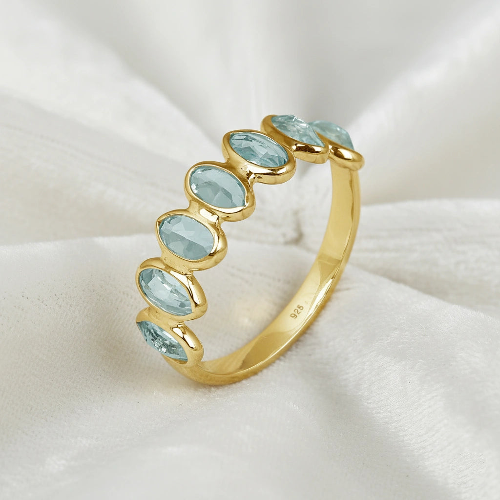 Gold-plated topaz ring band featuring seven bezel-set oval light blue gemstones on a white draped fabric side angle.