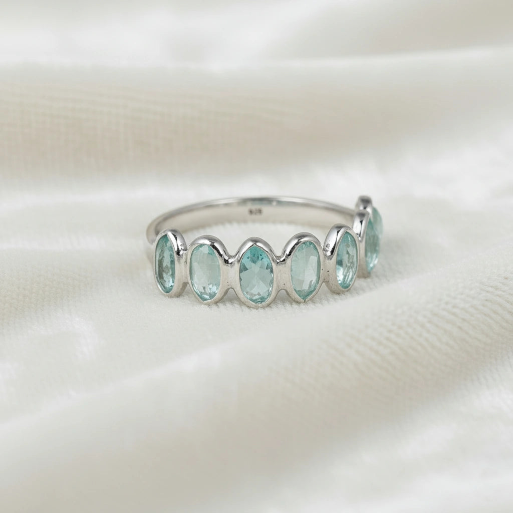 Sterling silver topaz ring band featuring seven bezel-set oval light blue gemstones on a soft white draped fabric front view.