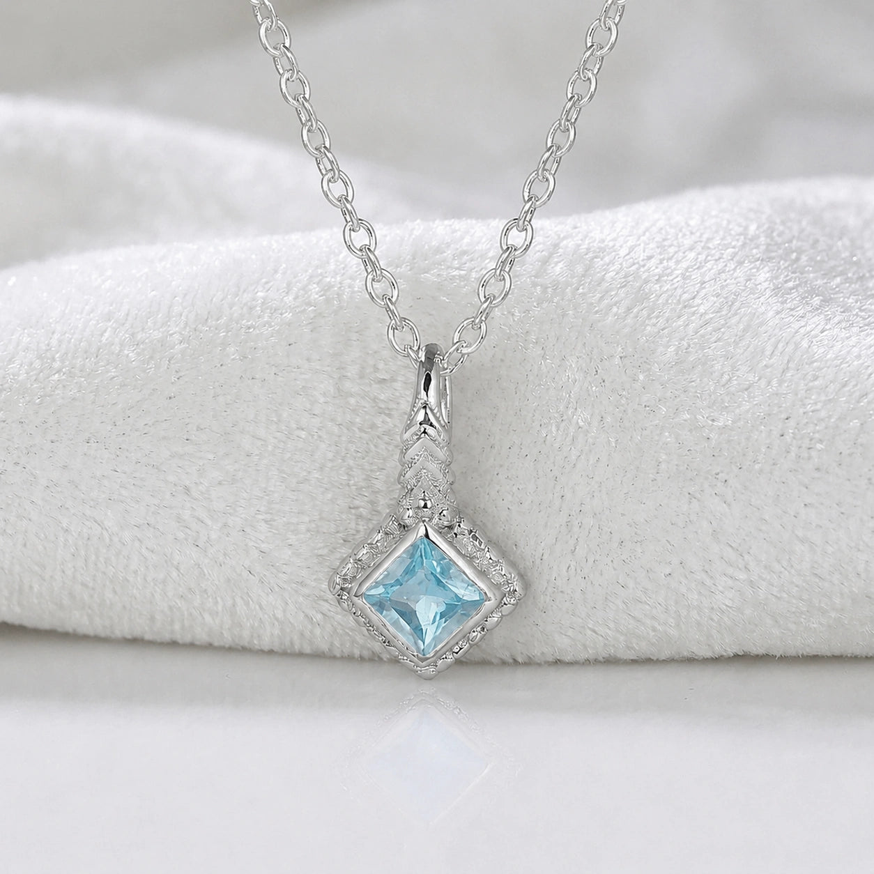 Birthstone_necklace_in_sterling_silver_with_square_cut_blue_topaz_pendant_in_front_angle_view_on_soft_white_fabric_background