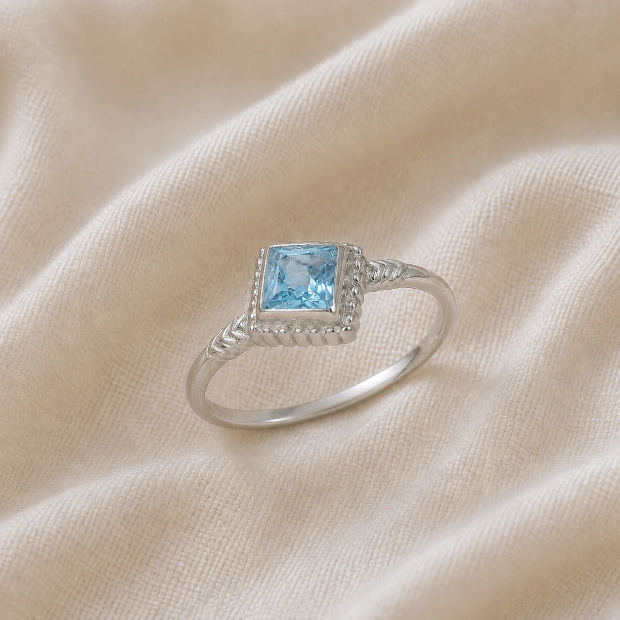 Princess_cut_ring_sterling_silver_with_square_blue_topaz_and_detailed_band_slightly_tilted_view_on_soft_fabric_background