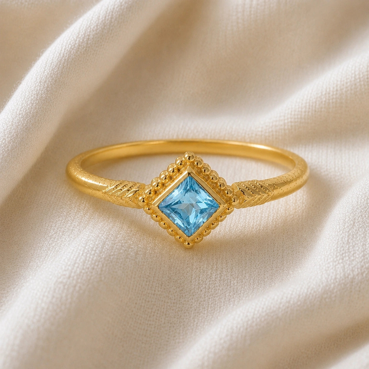 Princess_cut_ring_gold_plated_silver_with_square_blue_topaz_and_detailed_band_front_angle_view_on_soft_fabric_background
