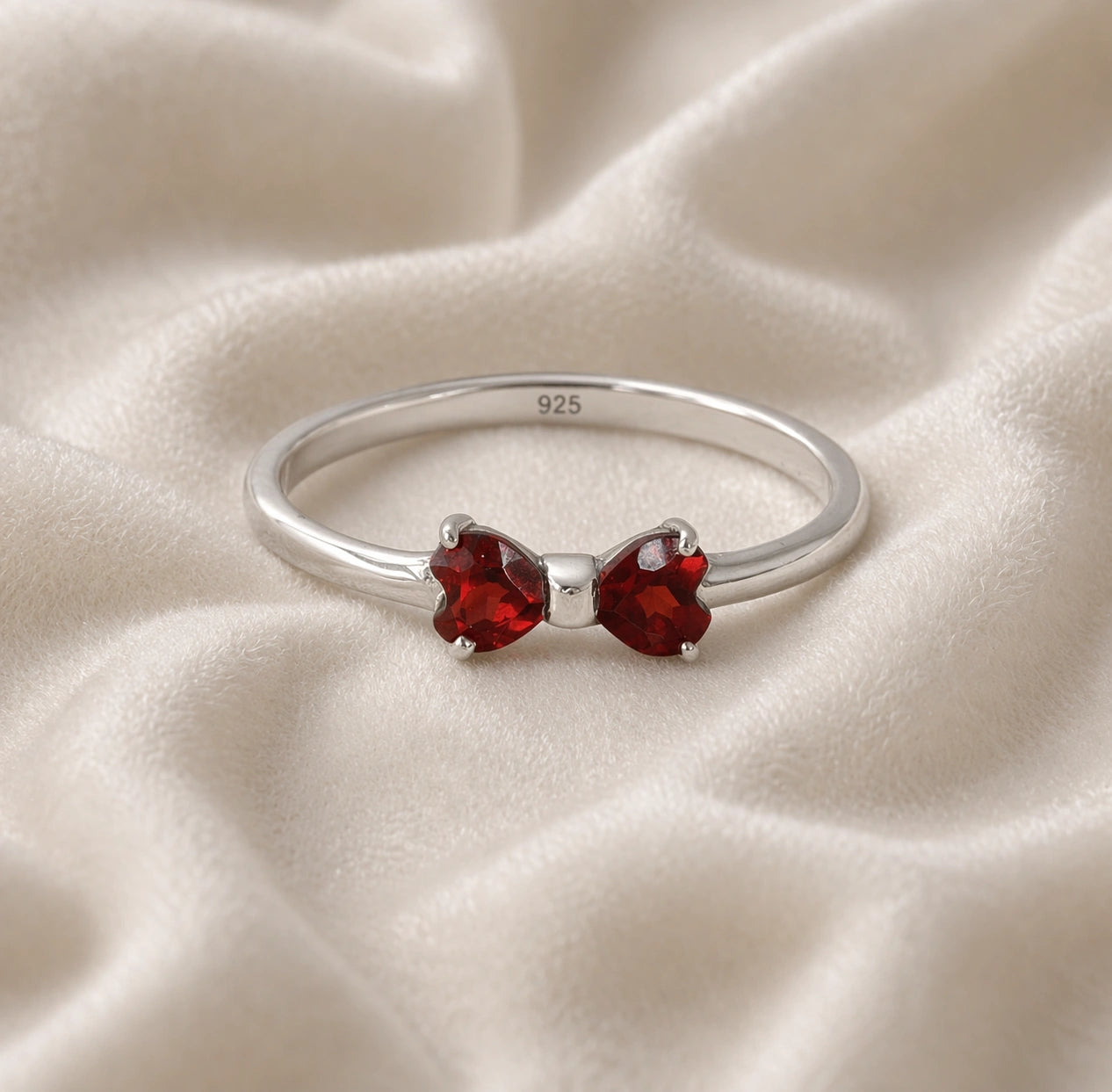 Bow_ring_close_up_front_angle_shot_sterling_silver_band_red_heart_garnet_gemstones_on_soft_off_white_fabric_background