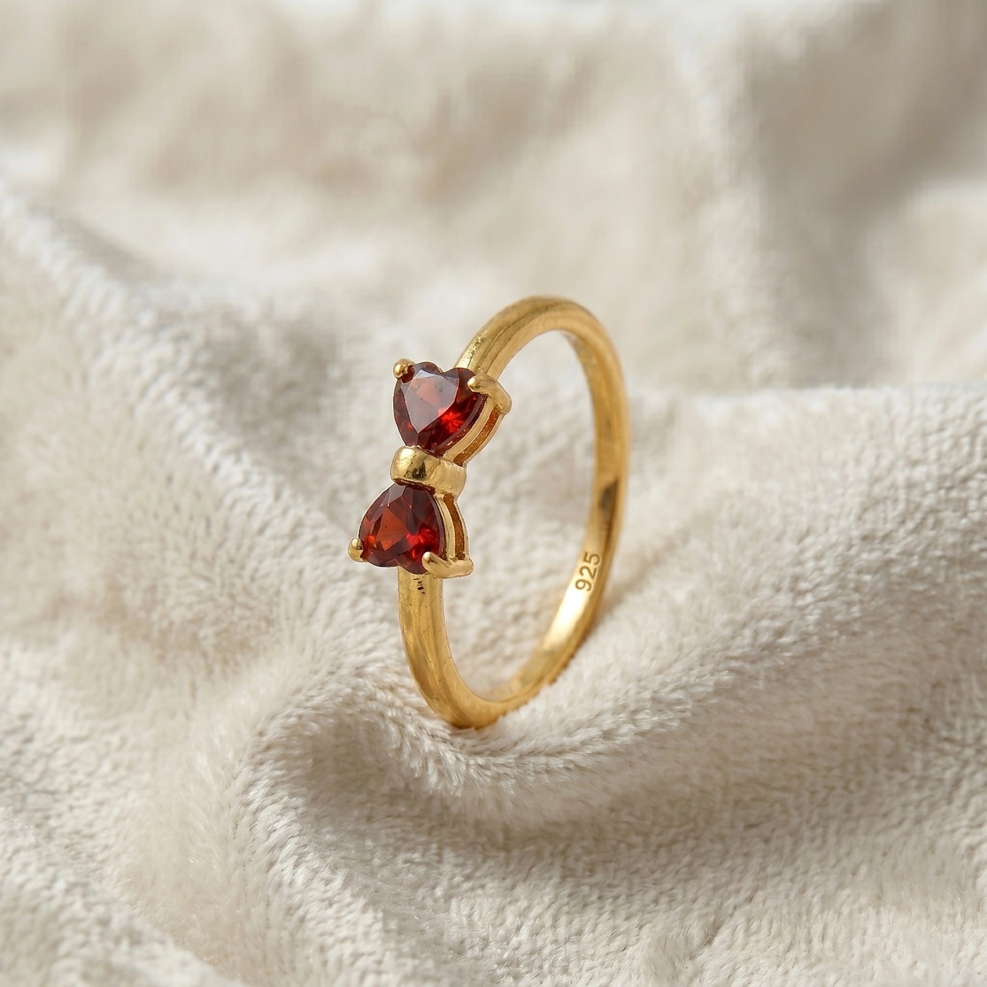 Bow_ring_close_up_slightly_angled_shot_gold_plated_silver_band_red_heart_garnet_gemstones_on_soft_off_white_fabric_background