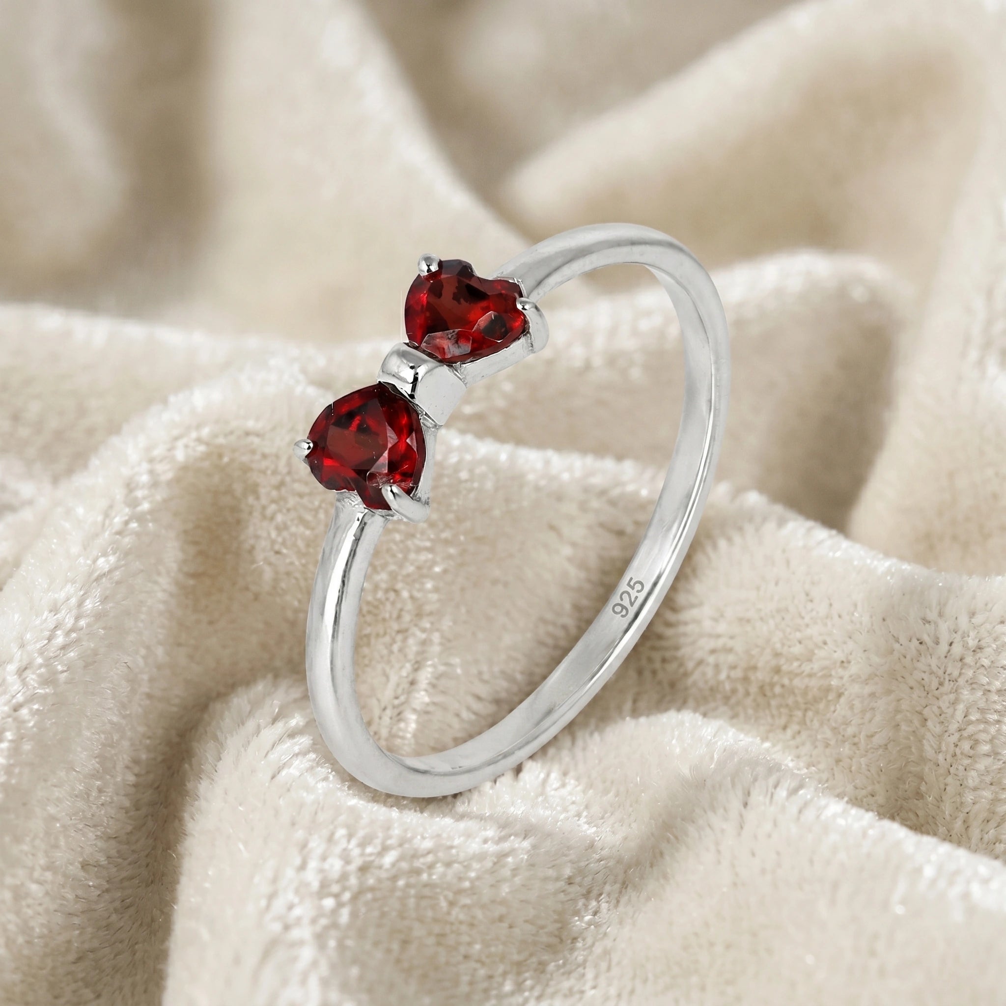 Bow_ring_close_up_slightly_angled_shot_sterling_silver_band_red_heart_garnet_gemstones_on_soft_off_white_fabric_background