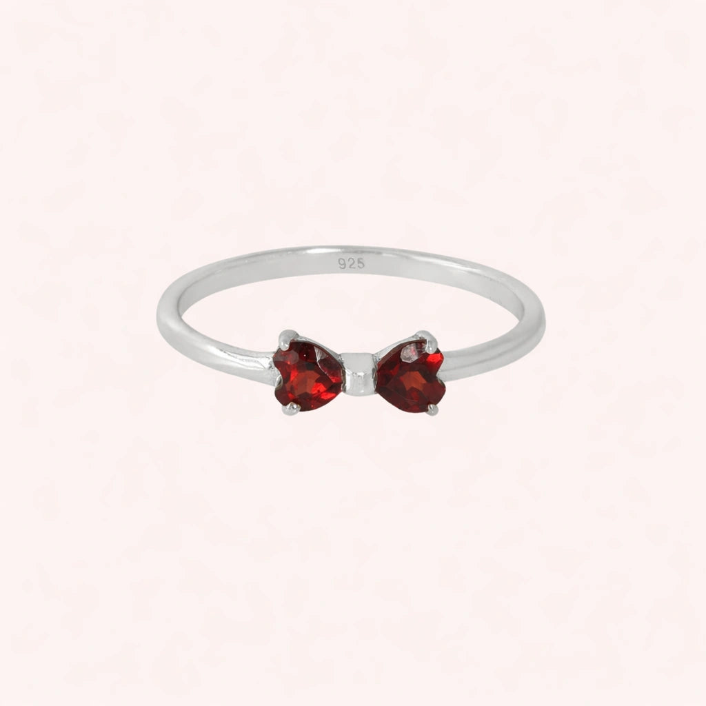 Bow_ring_close_up_front_angle_shot_sterling_silver_band_red_heart_garnet_gemstones_on_solid_pink_background