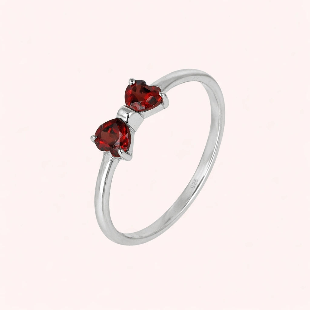 Bow_ring_close_up_slightly_angled_shot_sterling_silver_band_red_heart_garnet_gemstones_on_solid_pink_background