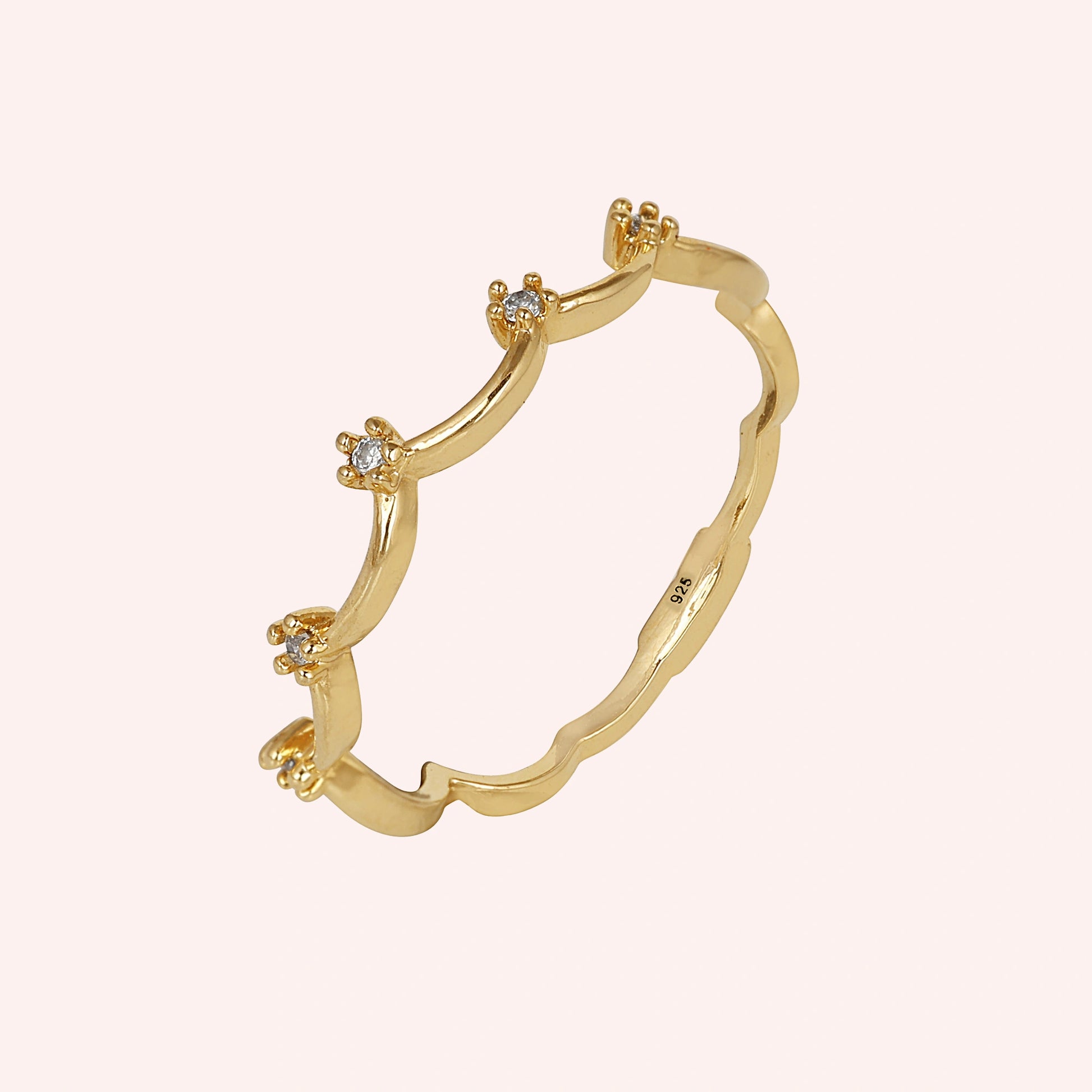 A gold plated crown ring with white topaz and a tiara ring design at a side angle over a pale pink plain background