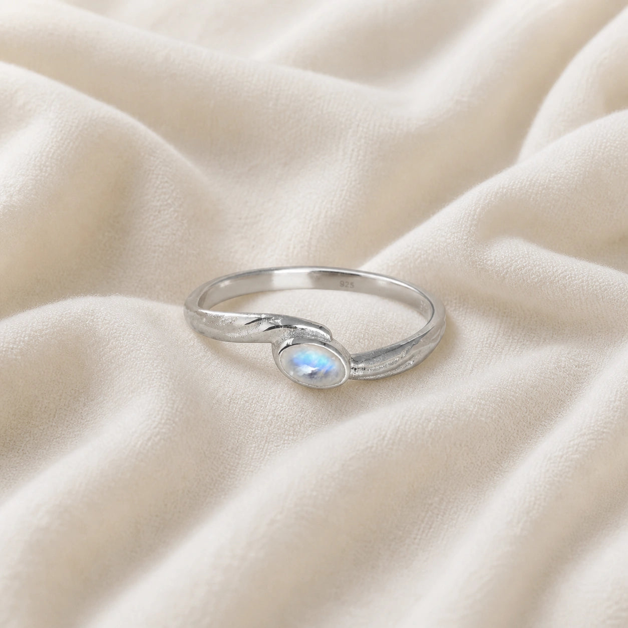 Cute_ring_sterling_silver_with_oval_moonstone_and_textured_wave_band_shown_at_front_angle_on_soft_fabric_background