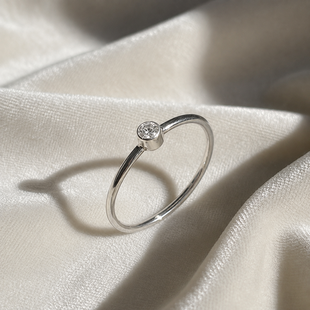 A silver birthstone ring featuring a diamond gemstone, styled as a delicate stacking ring on a soft white fabric background