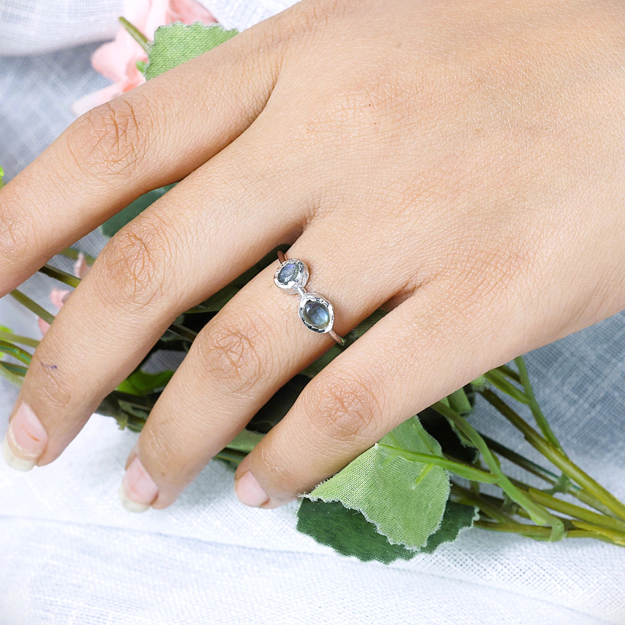 Double stone ring featuring two oval labradorite gems on a model's hand with flowers angled view