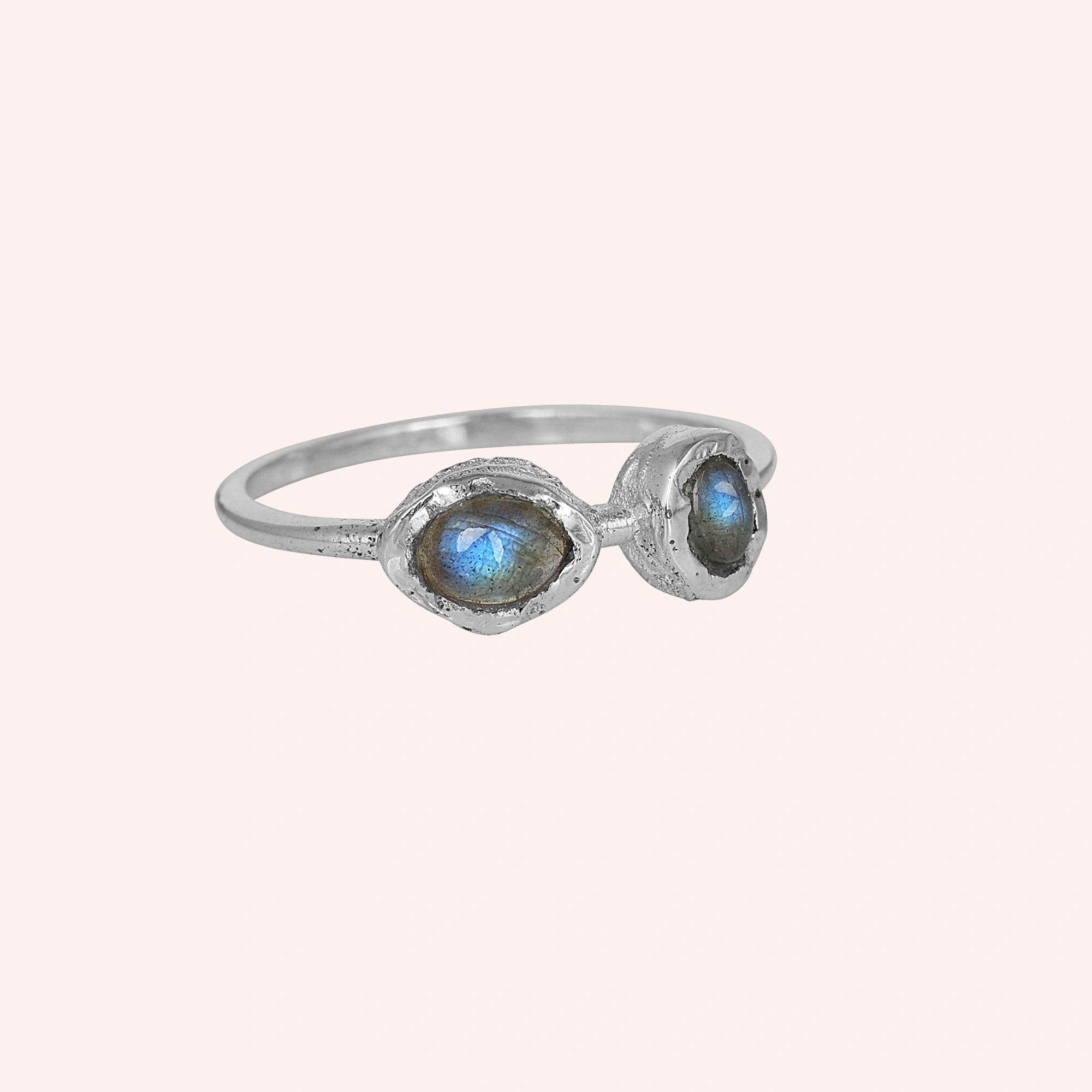 Double stone ring featuring two marquise labradorite gems in rugged silver settings on a pale pink background at a slight-right angle
