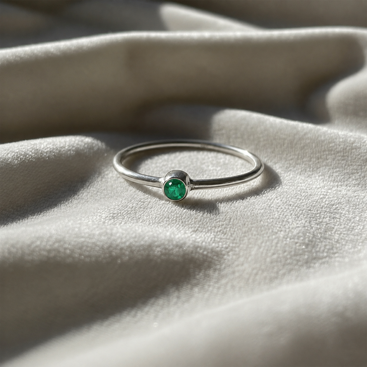 A silver birthstone ring featuring a emerald gemstone, styled as a delicate stacking ring on a soft white fabric background