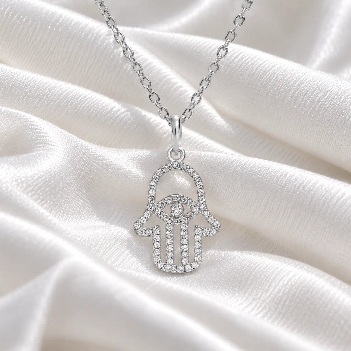 Evil_eye_necklace_in_sterling_silver_with_hamsa_hand_design_at_front_angle_view_on_soft_fabric_background