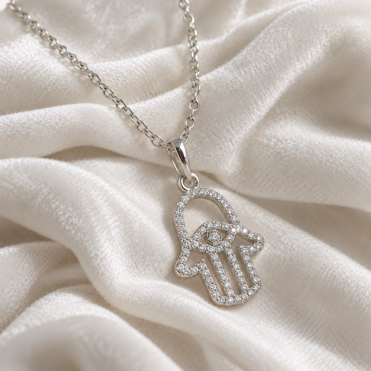 Evil_eye_necklace_in_sterling_silver_with_hamsa_hand_design_with_CZ_stones_at_side_angle_view_on_soft_fabric_background