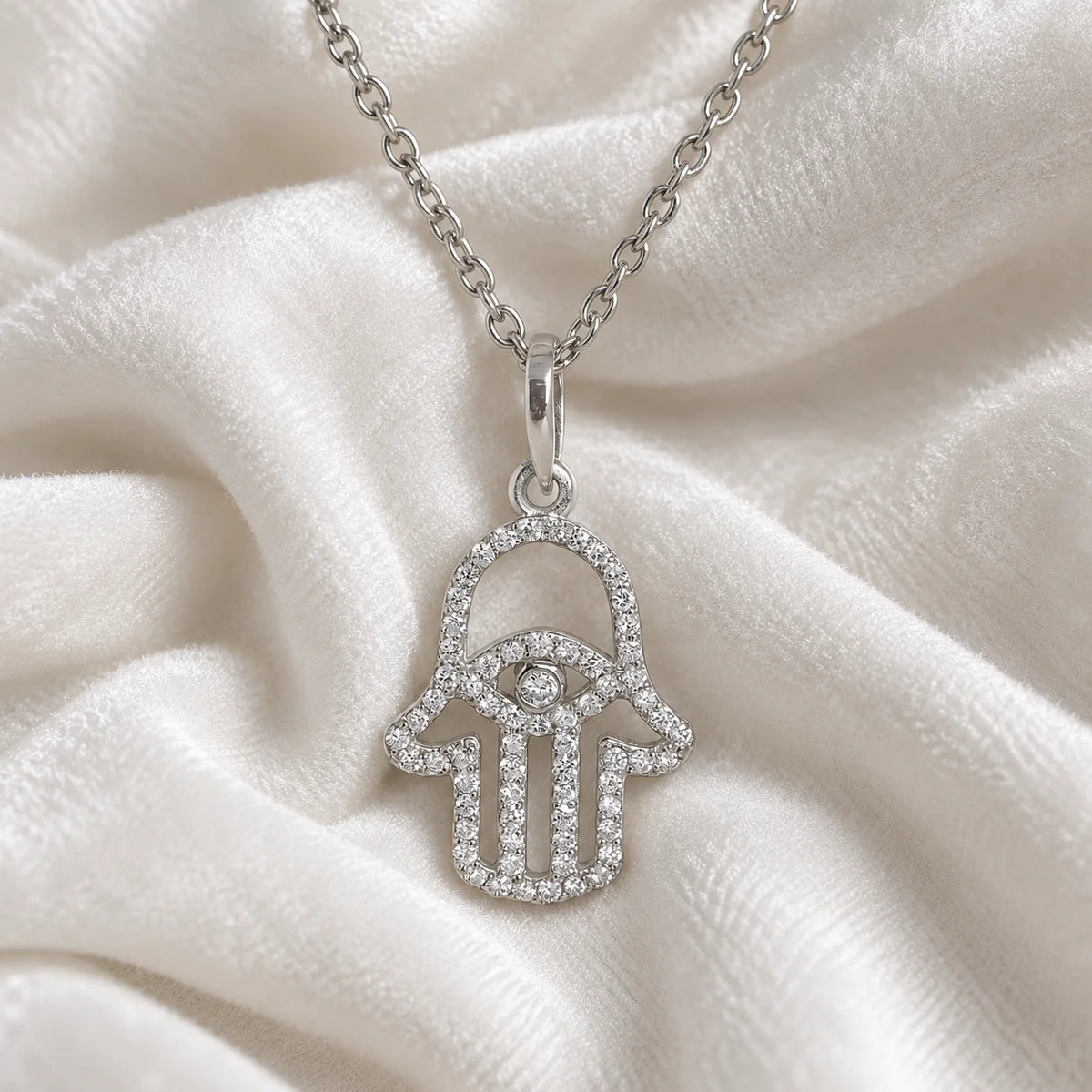 Evil_eye_necklace_in_sterling_silver_with_hamsa_hand_design_with_CZ_stones_at_front_angle_view_on_soft_fabric_background