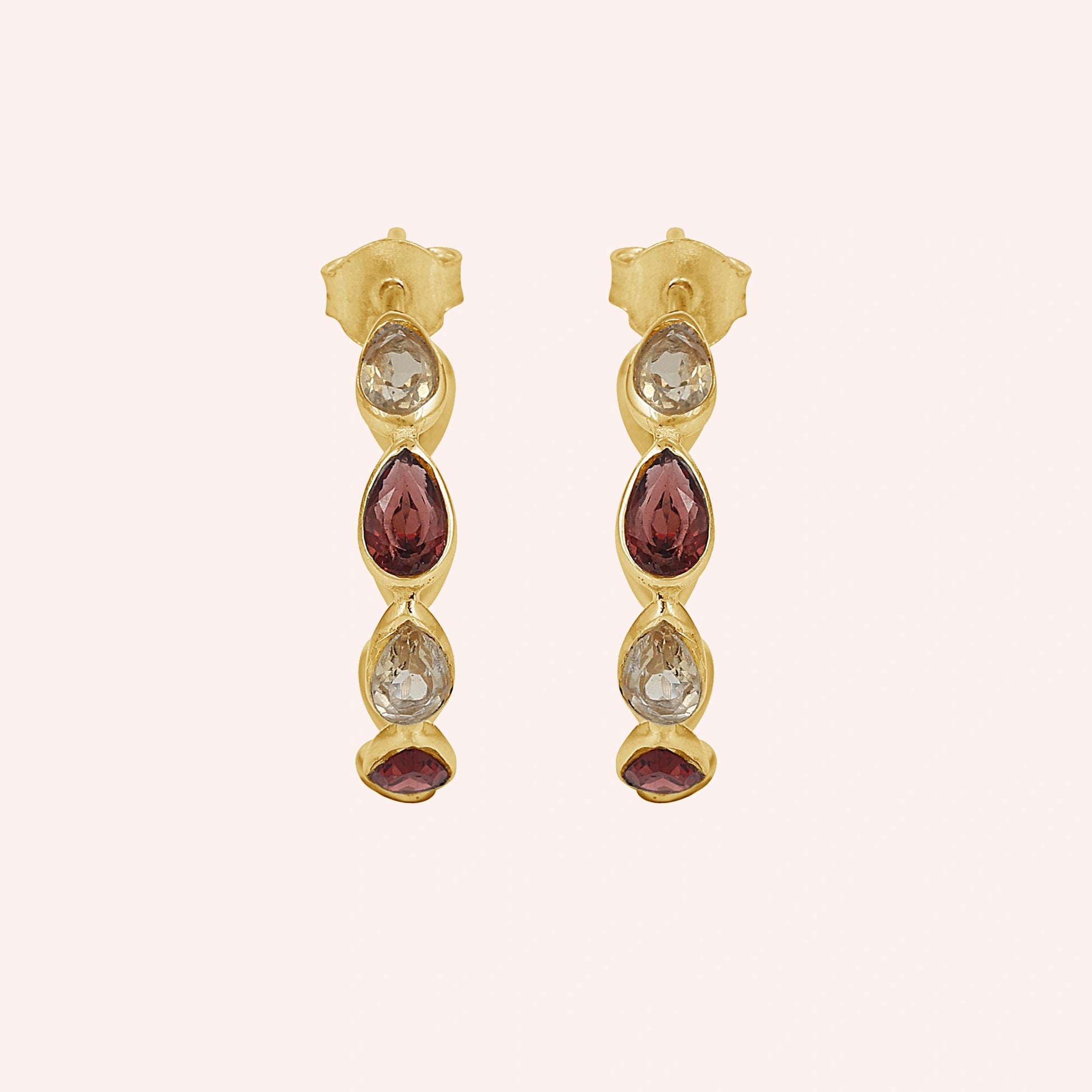 Gold plated hoop earrings with three light citrine and two garnet pear cut stones on a pink background top down angle shot