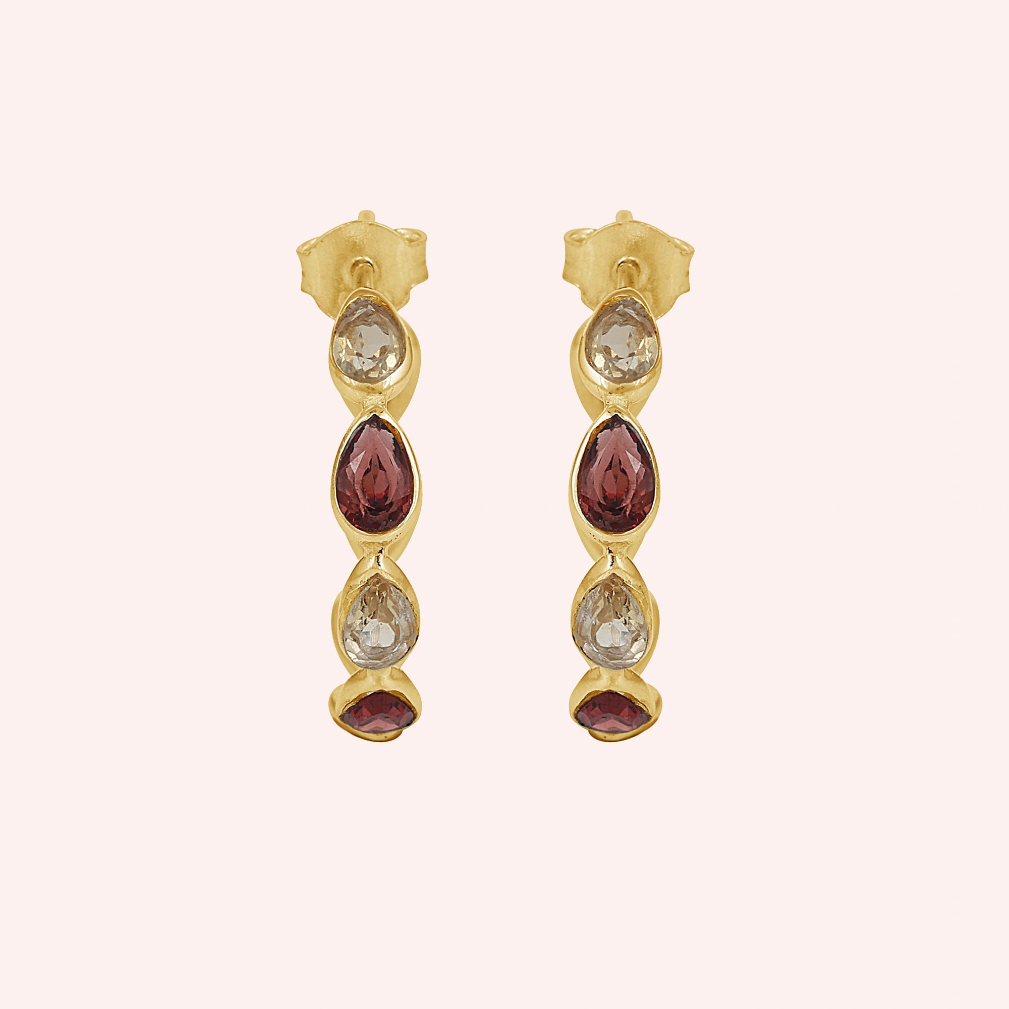 Gold plated hoop earrings with three light citrine and two garnet pear cut stones on a pink background top down angle shot