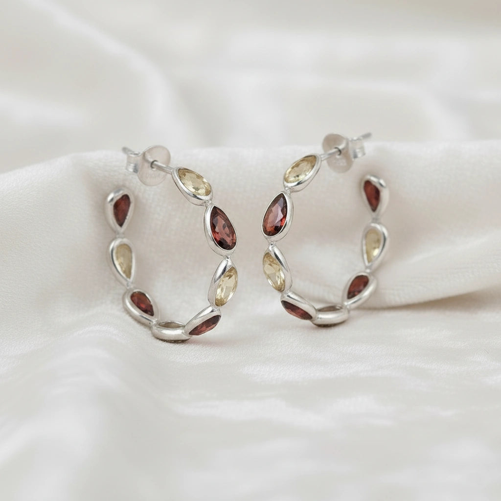 Silver hoop earrings featuring alternating citrine and garnet pear cut stones on an off white velvet fabric close up angled view shot