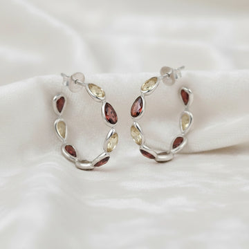 Silver hoop earrings featuring alternating citrine and garnet pear cut stones on an off white velvet fabric close up angled view shot