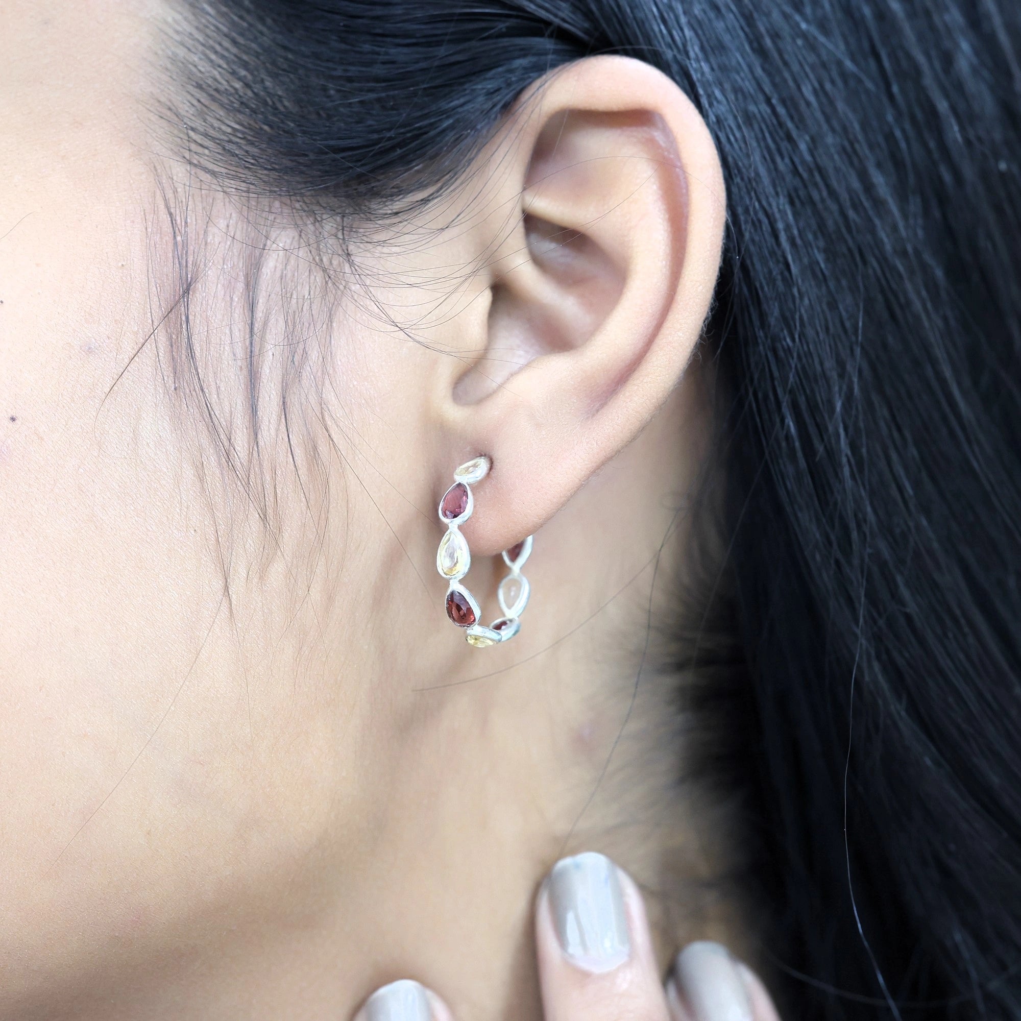 Side angle view of silver hoop earrings featuring red and yellow stones worn on the ear with the hand touching the neck view