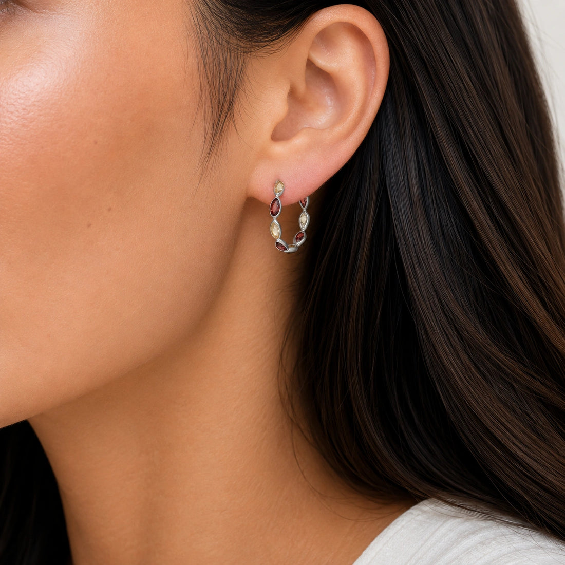 Silver hoop earrings featuring alternating citrine and garnet pear cut stones on an off white velvet fabric close up angled view shot