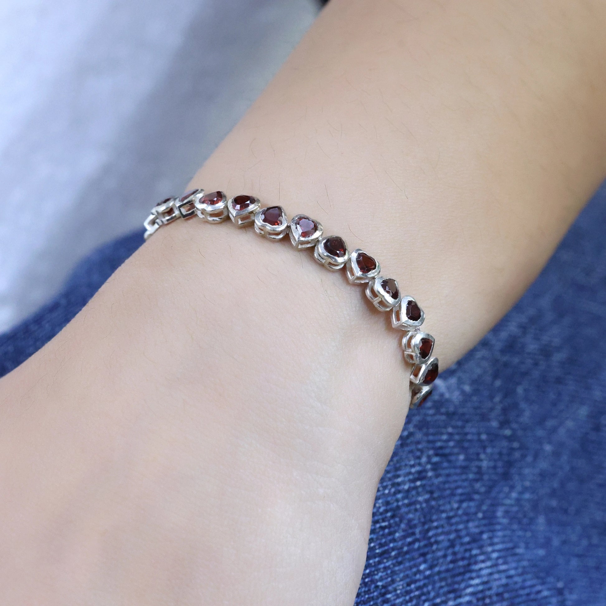 Angled view of sterling silver garnet bracelet with red heart stones worn on a wrist over blue denim jeans background texture