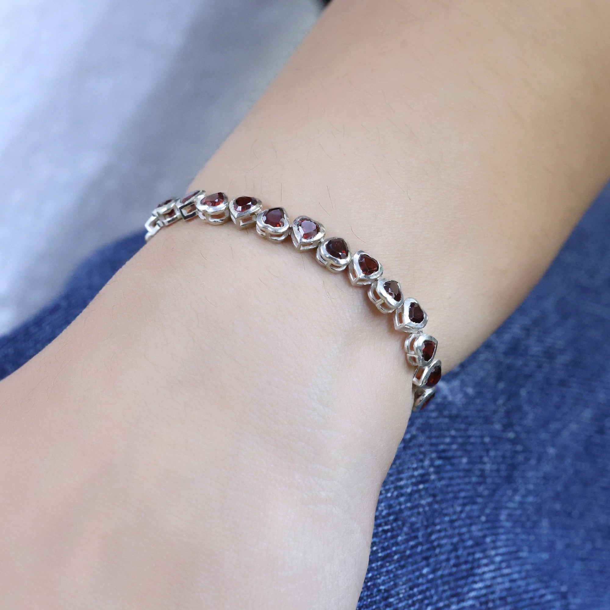 Angled view of sterling silver garnet bracelet with red heart stones worn on a wrist over blue denim jeans background texture