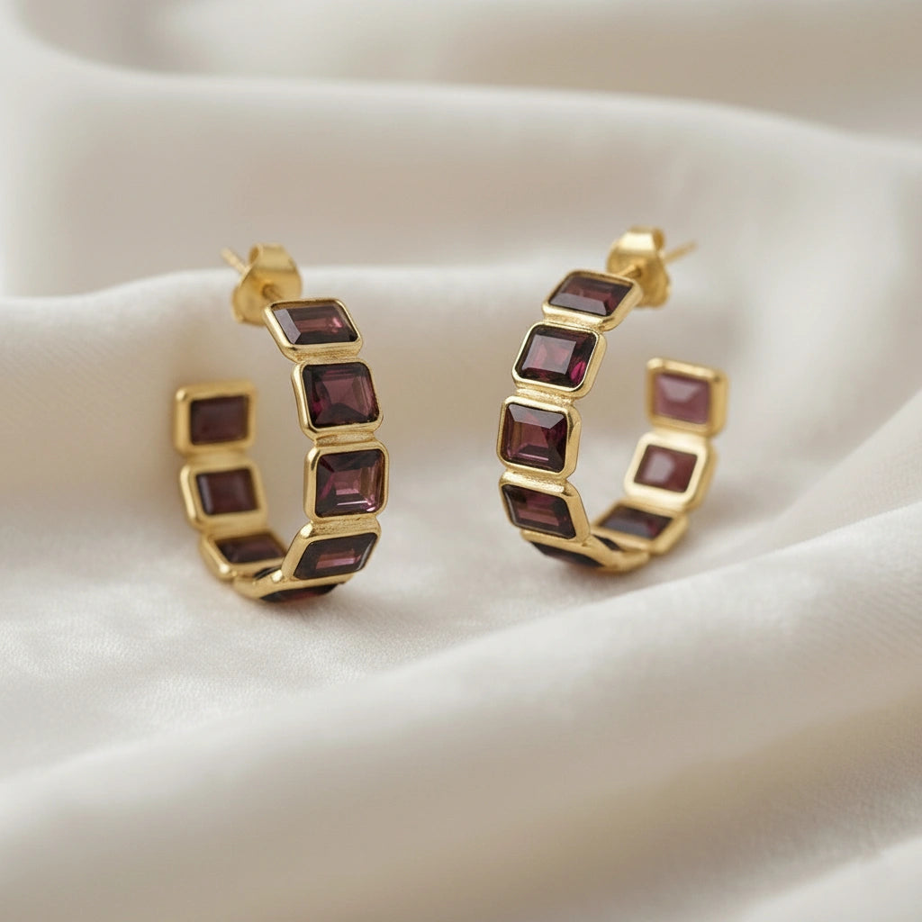 Garnet hoop earrings with five dark red square stones in gold setting on wrinkled off white velvet fabric close up angled shot