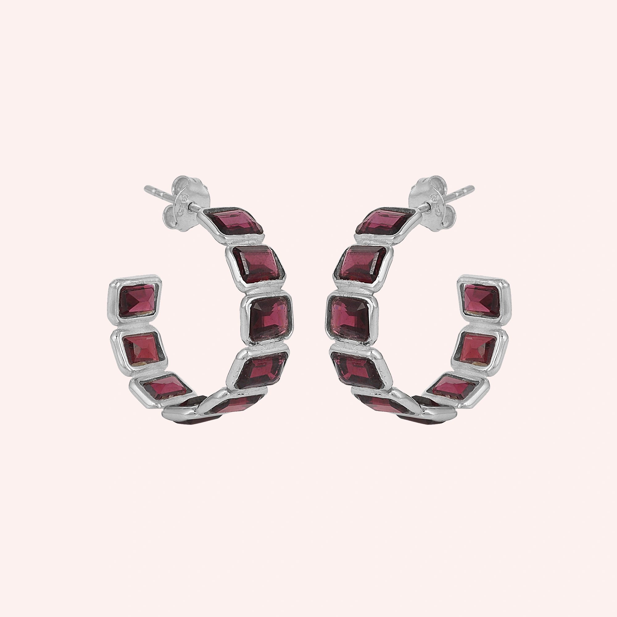 Pair of garnet hoop earrings featuring five dark red square stones in a silver setting on a pink background slightly angled view