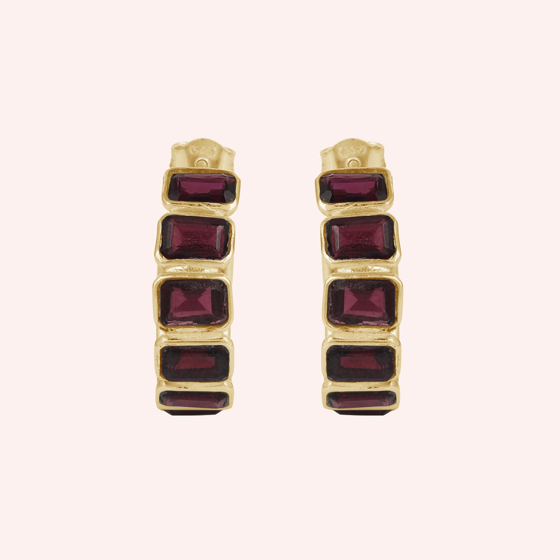 Pair of garnet hoop earrings featuring four dark red rectangular stones in a gold setting on a pink background top down angle