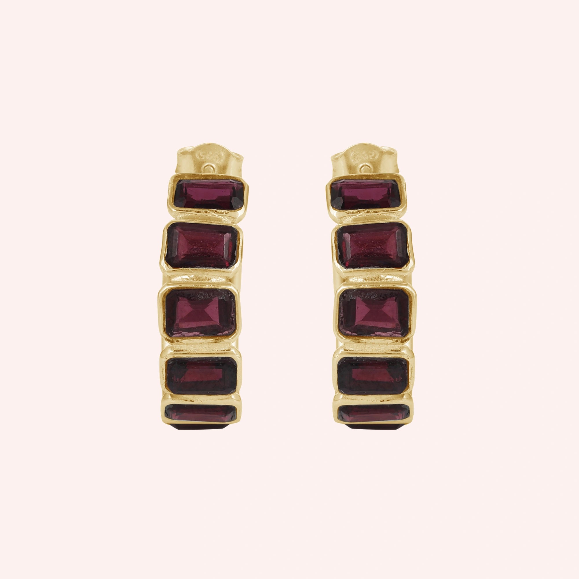 Pair of garnet hoop earrings featuring four dark red rectangular stones in a gold setting on a pink background top down angle