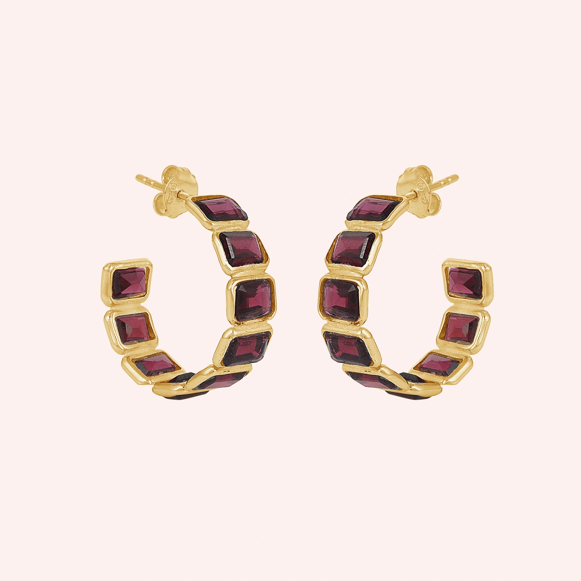 Pair of garnet hoop earrings featuring five dark red square stones in a gold setting on a pink background slightly angled view