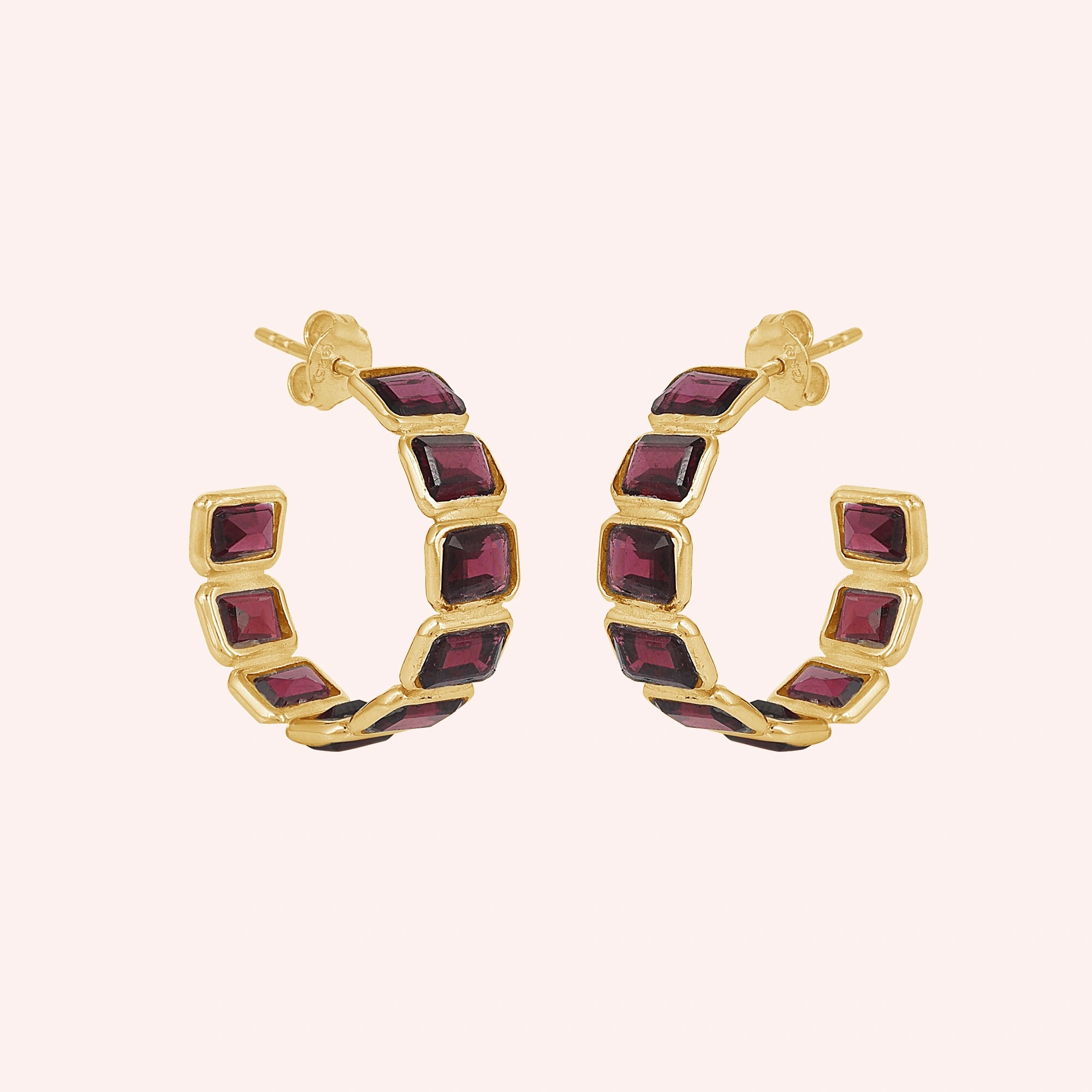 Pair of garnet hoop earrings featuring five dark red square stones in a gold setting on a pink background slightly angled view