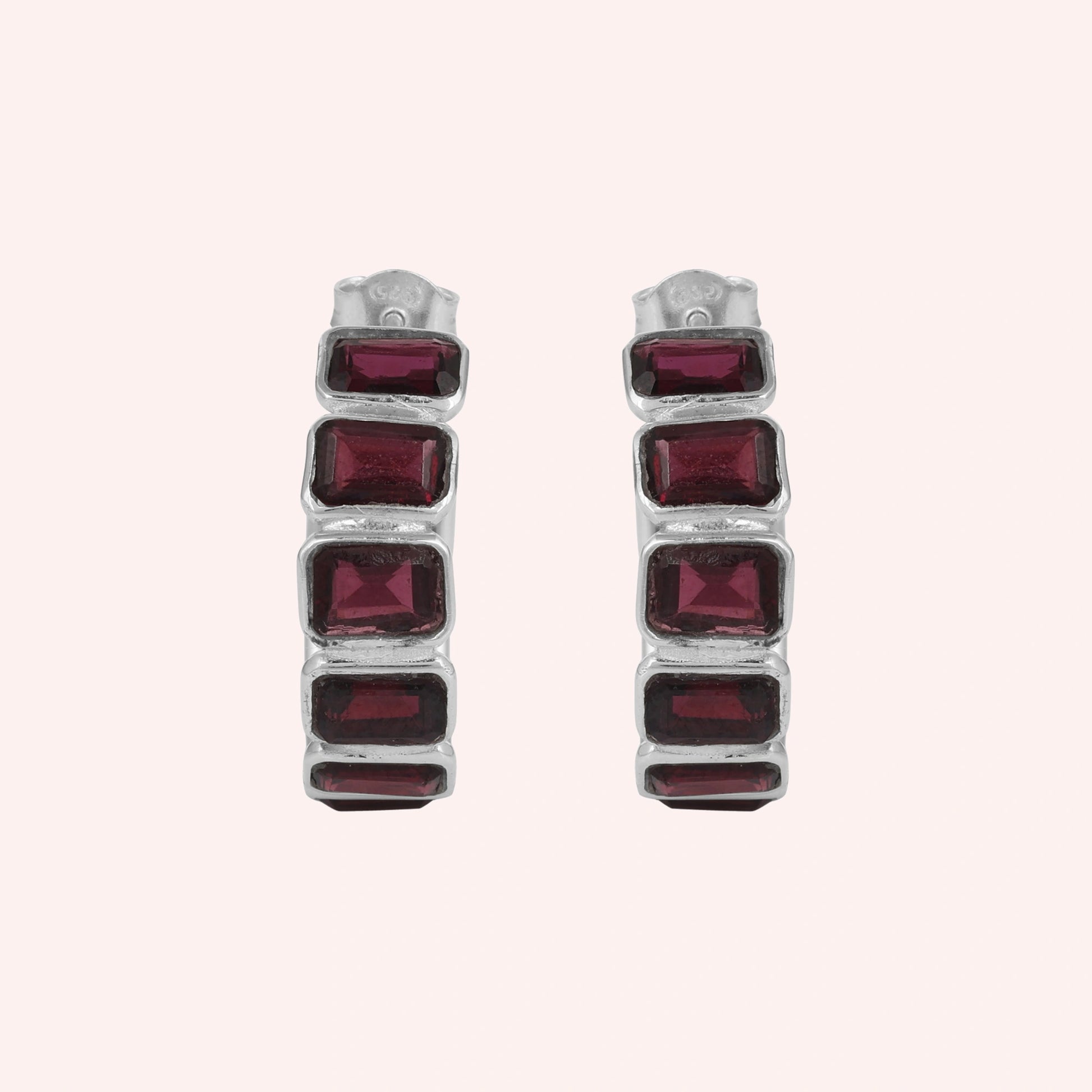 Pair of garnet hoop earrings featuring four dark red rectangular stones in a silver setting on a pink background top down angle