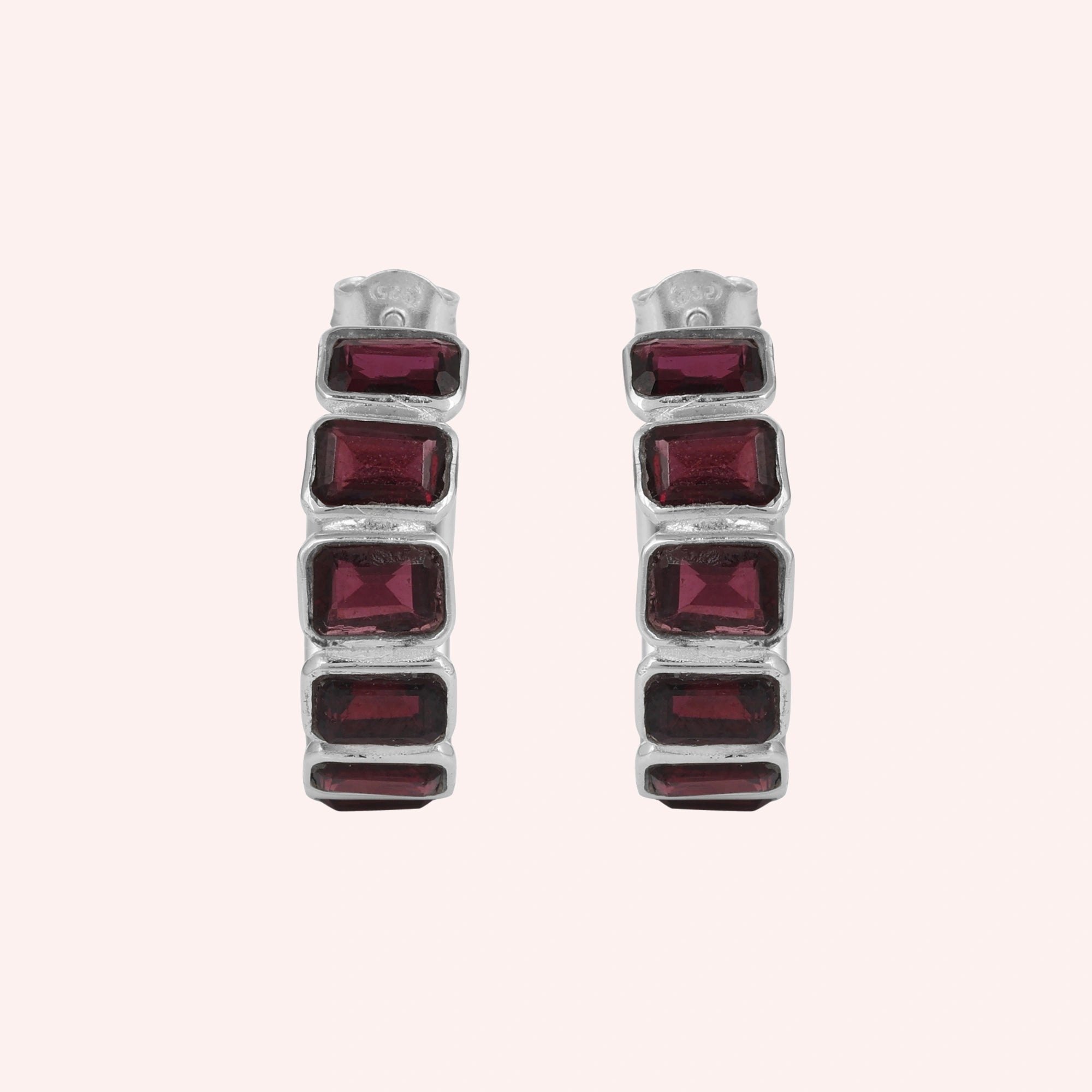 Pair of garnet hoop earrings featuring four dark red rectangular stones in a silver setting on a pink background top down angle