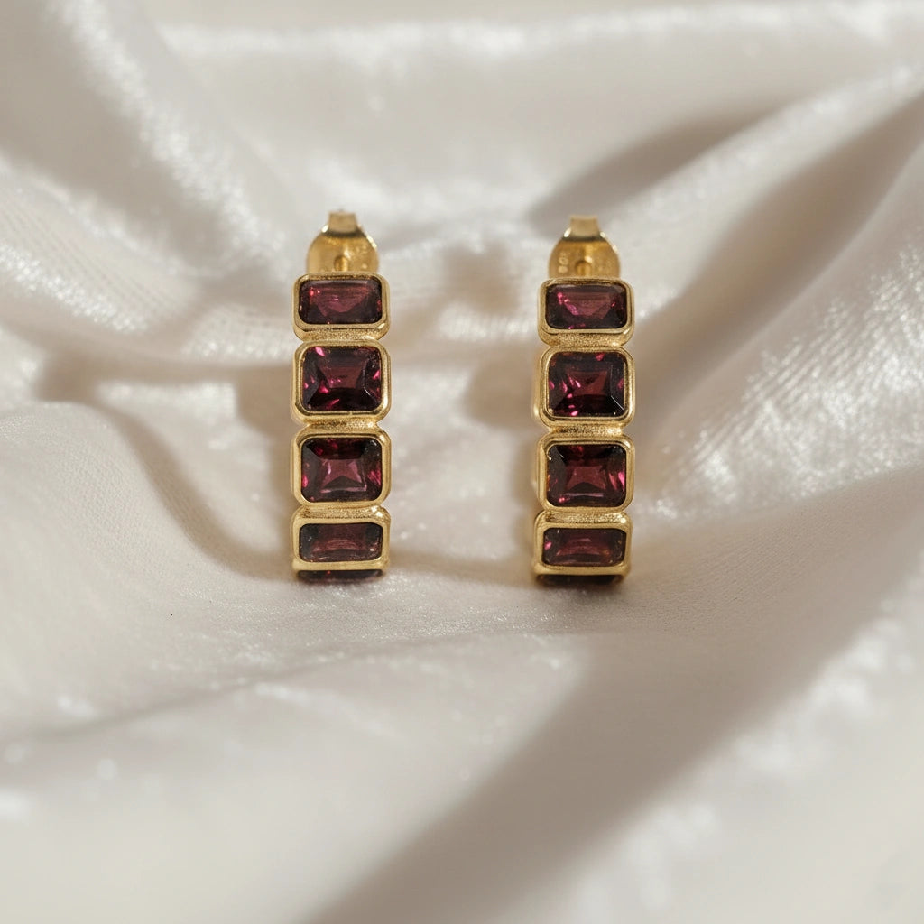 Garnet hoop earrings with four dark red rectangular stones in gold setting on wrinkled off white velvet fabric close up shot