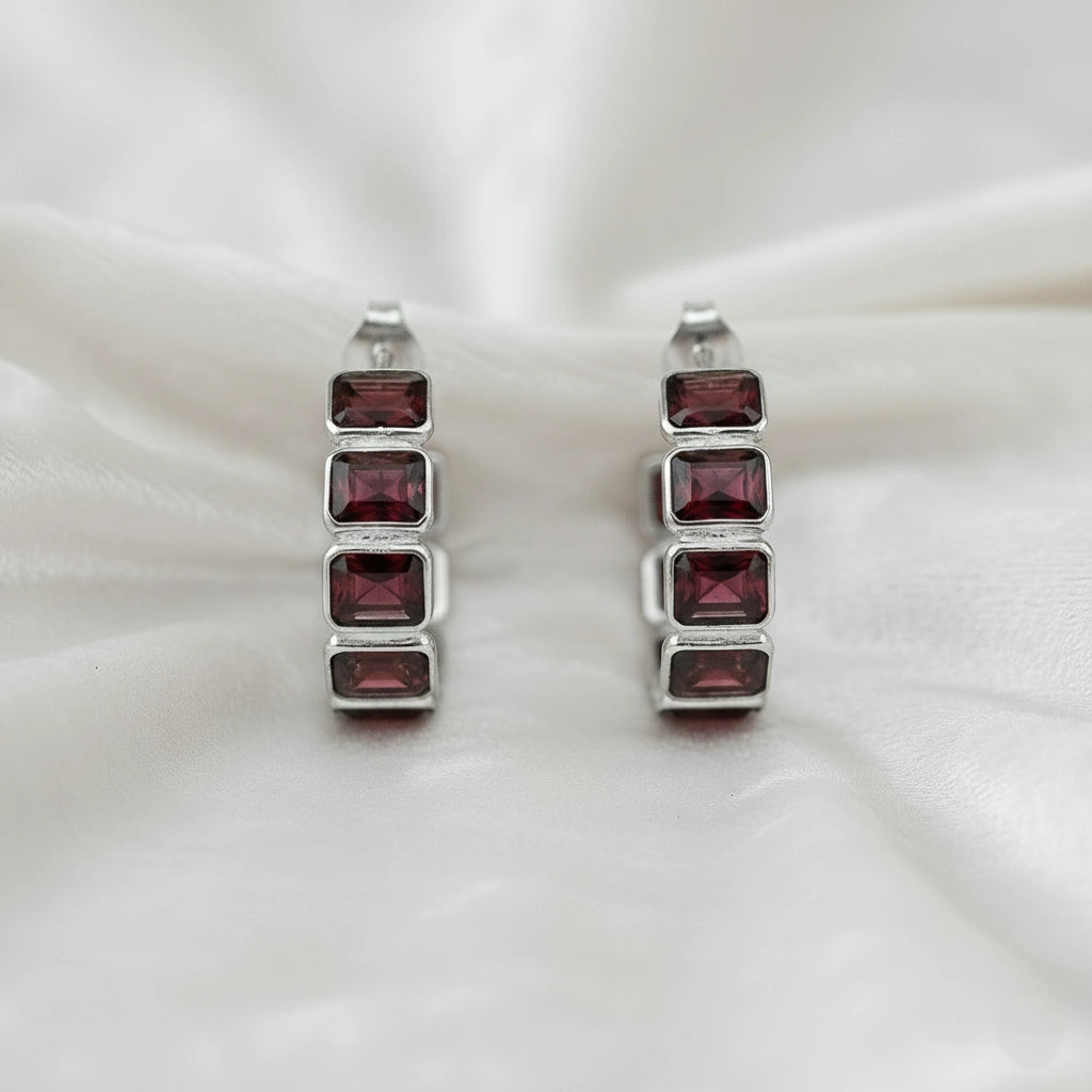 Pair of silver garnet hoop earrings featuring four dark red rectangular stones on a wrinkled off white velvet fabric top down angle shot