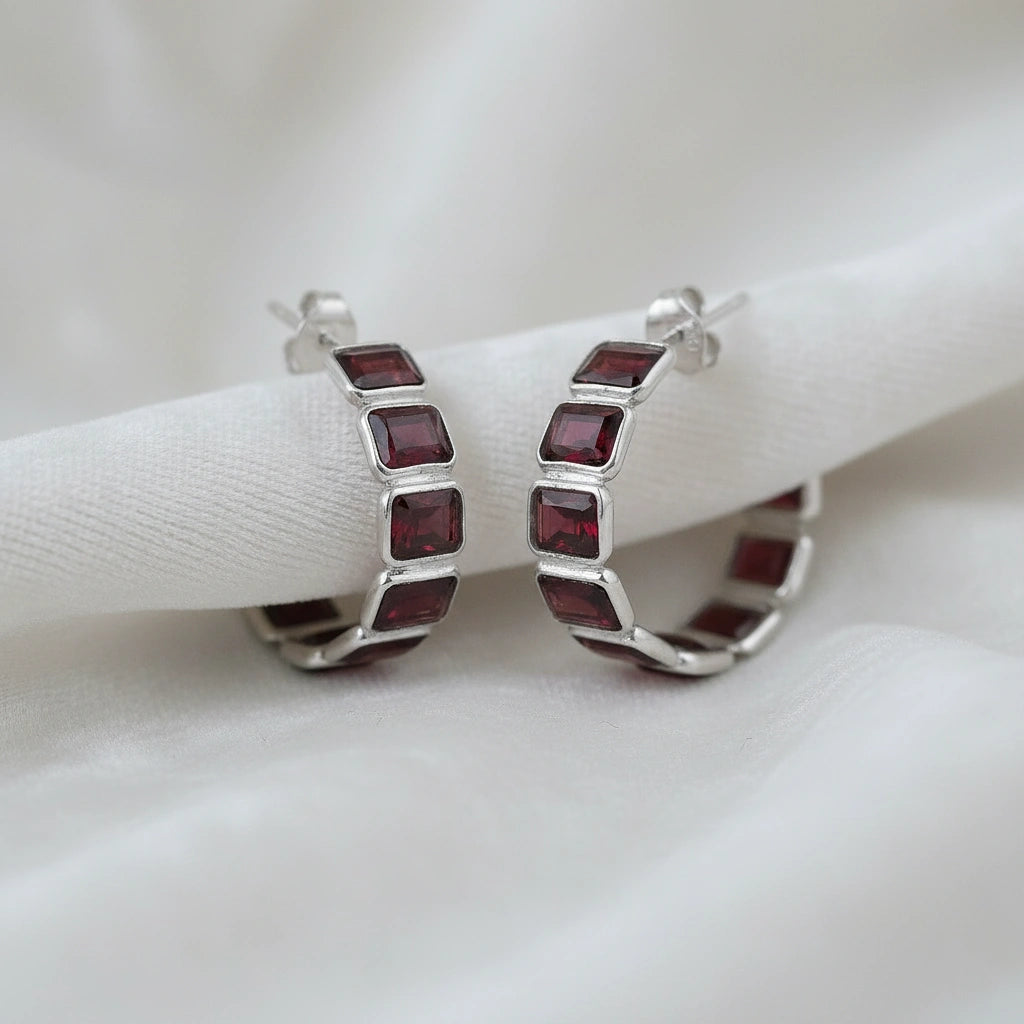 Pair of silver garnet hoop earrings featuring five dark red square stones on an off white velvet fabric close up angle shot