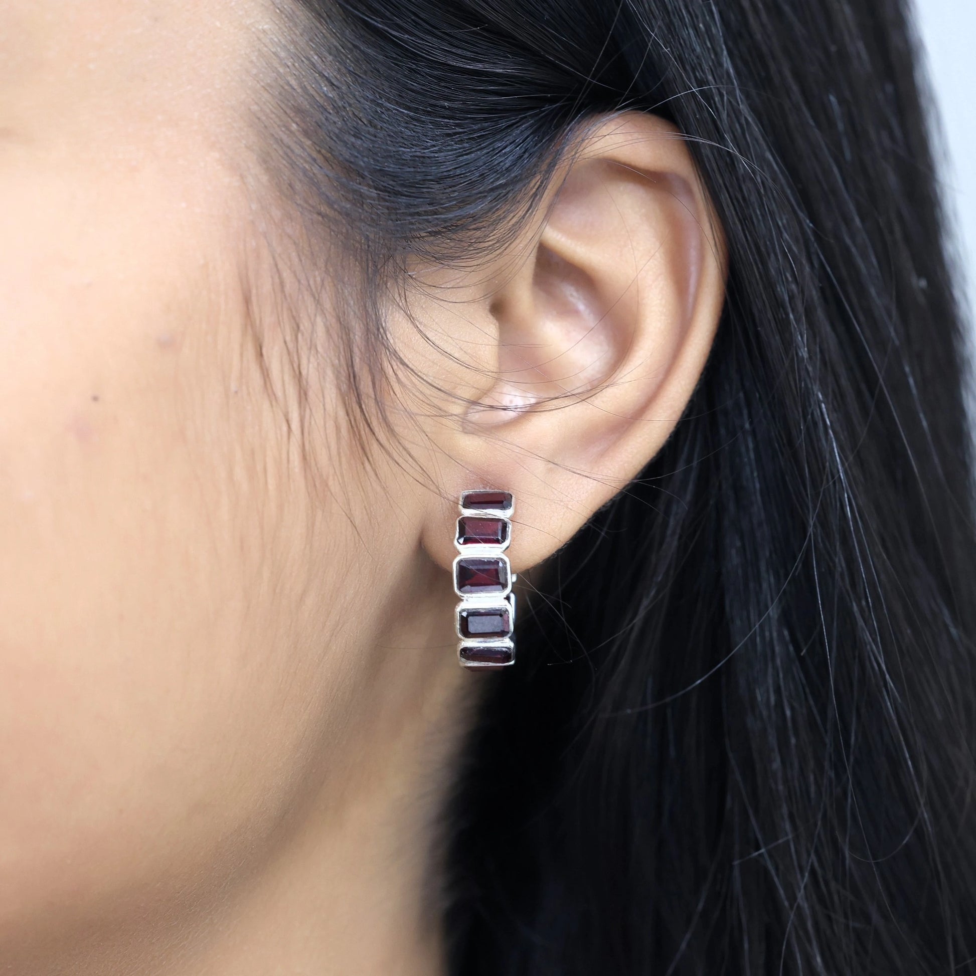 Side angle view of silver garnet hoop earrings with red garnet baguette stones worn on ear with background texture