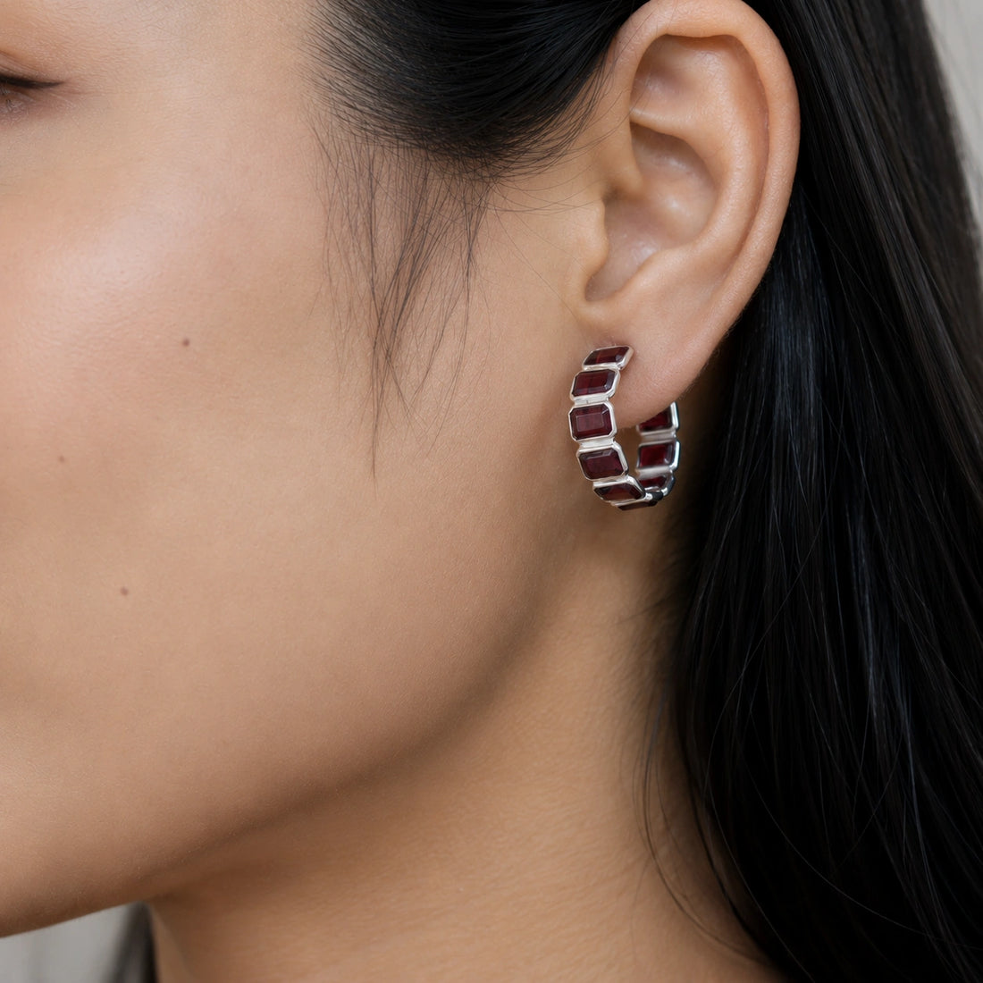 Pair of silver garnet hoop earrings featuring five dark red square stones on an off white velvet fabric close up angle shot