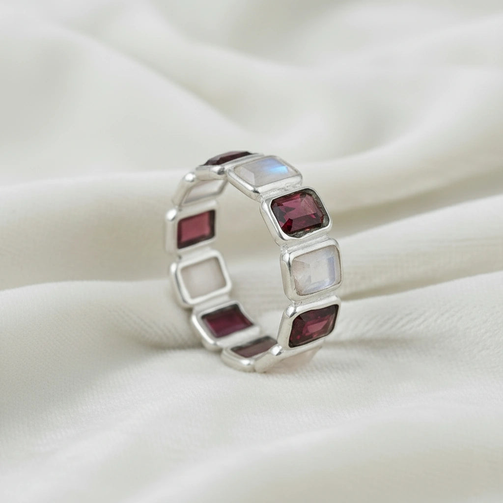 Silver Garnet ring band with alternating rectangular moonstone and ruby colored gems on a draped white fabric slight side angle.