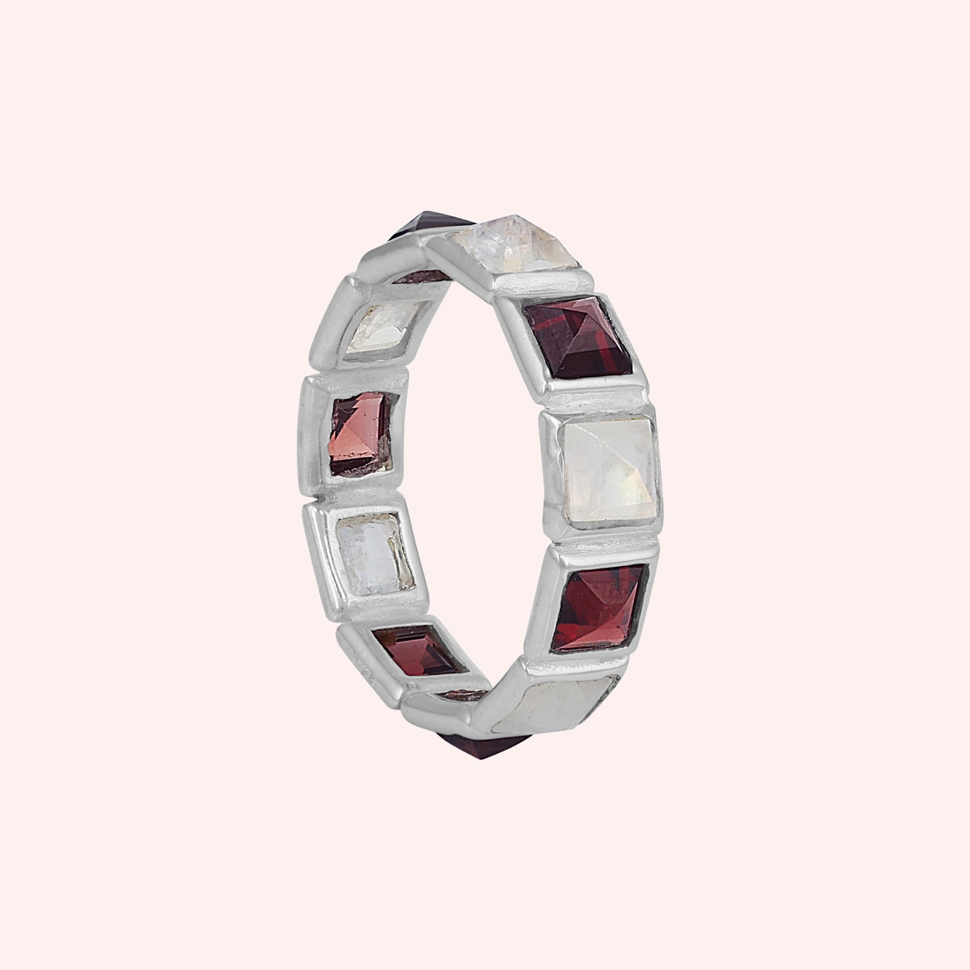 Angled shot of a silver moonstone ring on a light pink background with alternating square moonstone and garnet stones