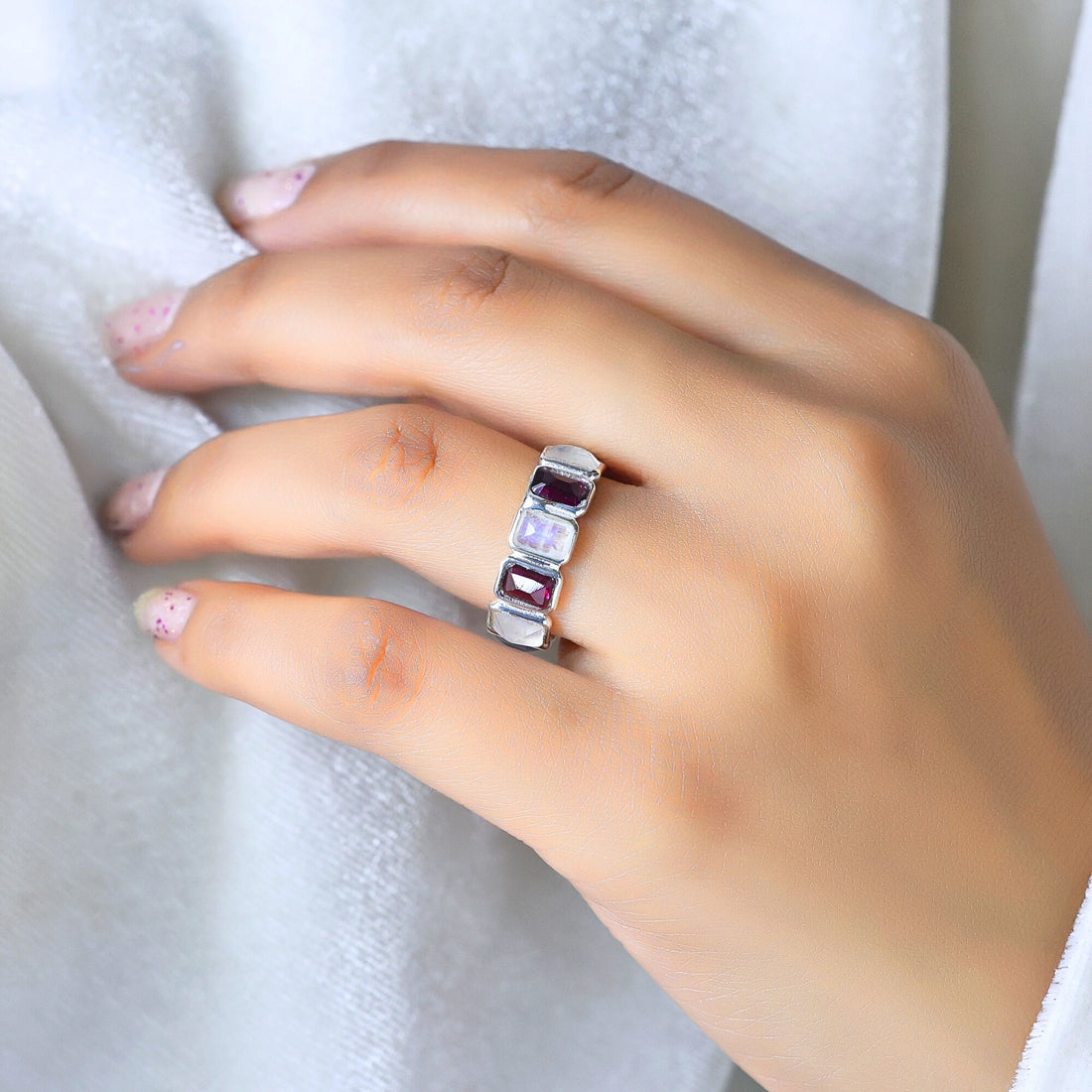 Silver Garnet ring band with alternating rectangular moonstone and ruby colored gems on a draped white fabric slight side angle.