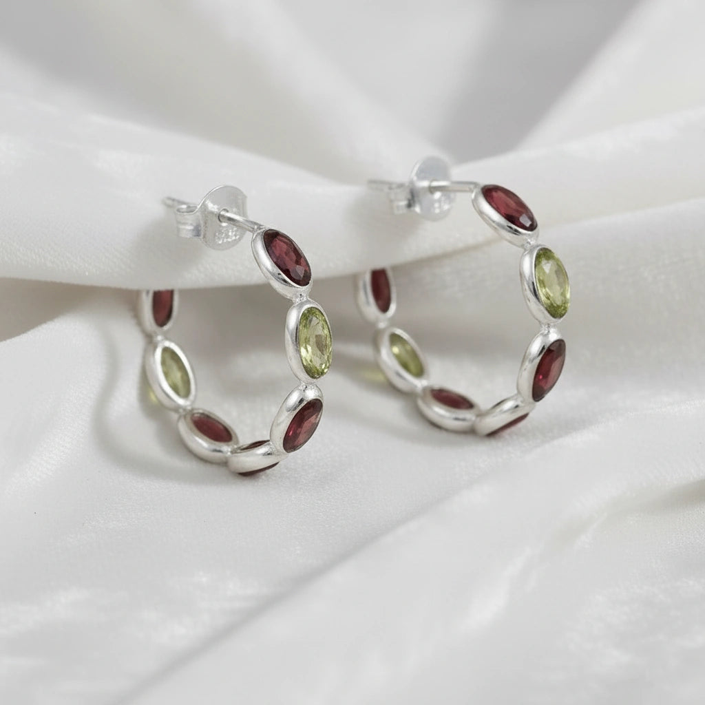 Silver hoop earrings featuring oval red garnet and green peridot stones on an off white velvet fabric close up angled view shot