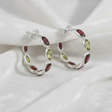 Silver hoop earrings featuring oval red garnet and green peridot stones on an off white velvet fabric close up angled view shot