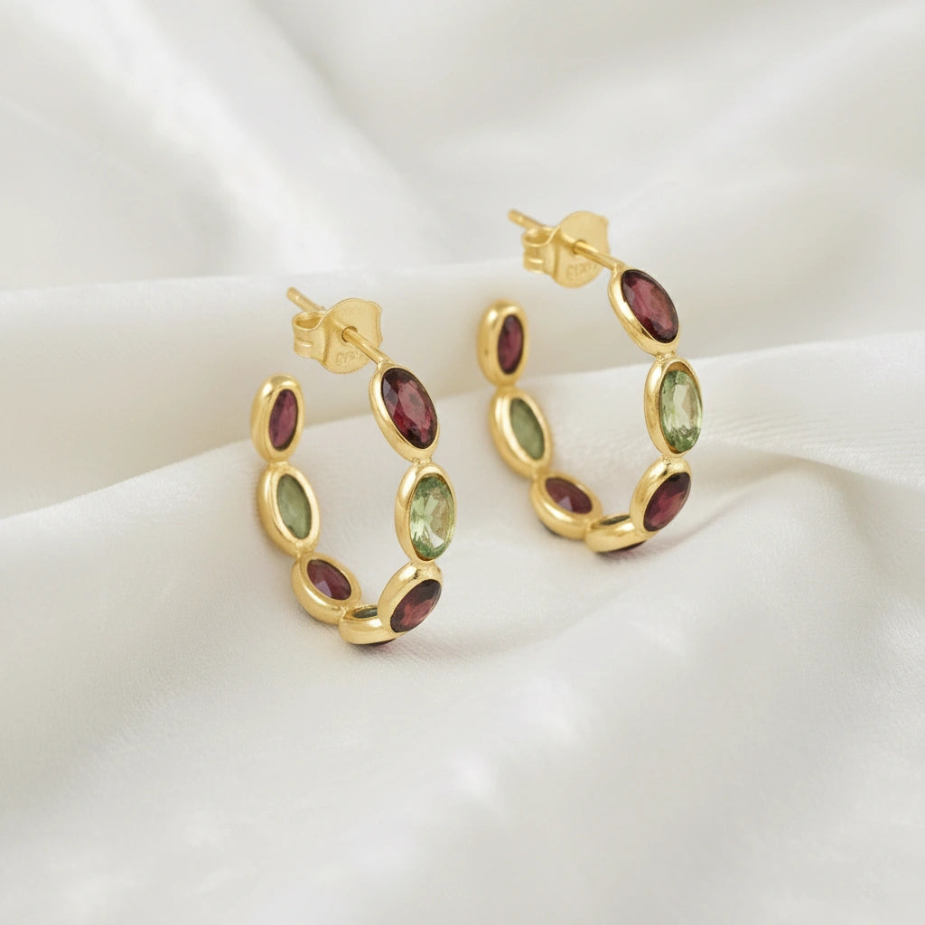 Gold plated hoop earrings featuring oval red garnet and green peridot stones on an off white velvet fabric close up angled view shot
